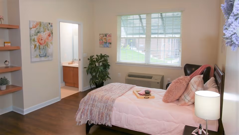 A cozy bedroom with a neatly made bed featuring pink bedding and pillows. There is a bedside table with a lamp and a small plant. A large window with blinds lets in natural light, and a green plant is placed near the doorway leading to a bathroom with a sink and mirror. The room has wooden floors and light-colored walls decorated with floral paintings.