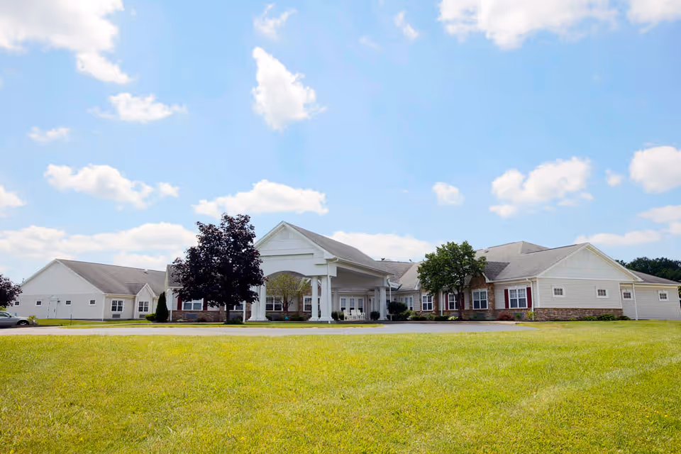 Exterior view of a single-story senior living facility building with a large covered entrance, surrounded by green grass and trees under a partly cloudy blue sky.