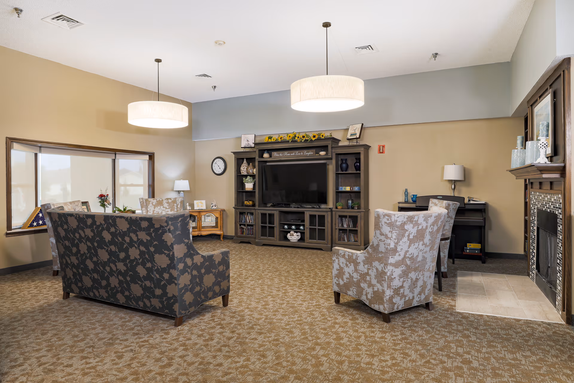 A spacious living room with patterned armchairs and a sofa arranged around a large entertainment center with a flat-screen TV. The room features beige walls, carpeted flooring, two hanging ceiling lights, a fireplace with decorative vases on the mantel, a clock on the wall, and a large window with a view outside. There is also a small desk with a lamp in the corner.