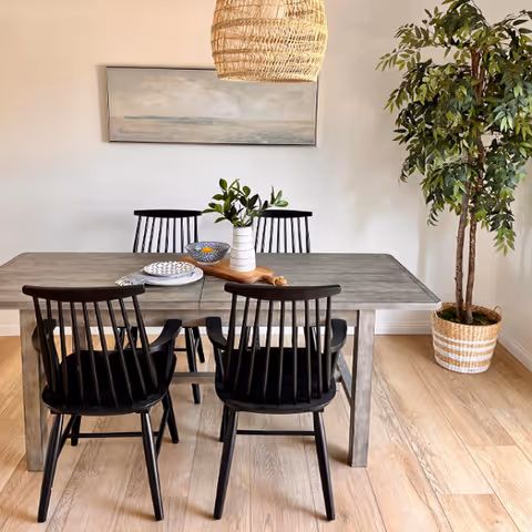 Modern dining room with a wooden table, four black chairs, a woven pendant light, wall art, and a potted plant.