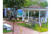 Outdoor garden area with a white gazebo, benches, a brick pathway, and green grass surrounded by trees and plants.
