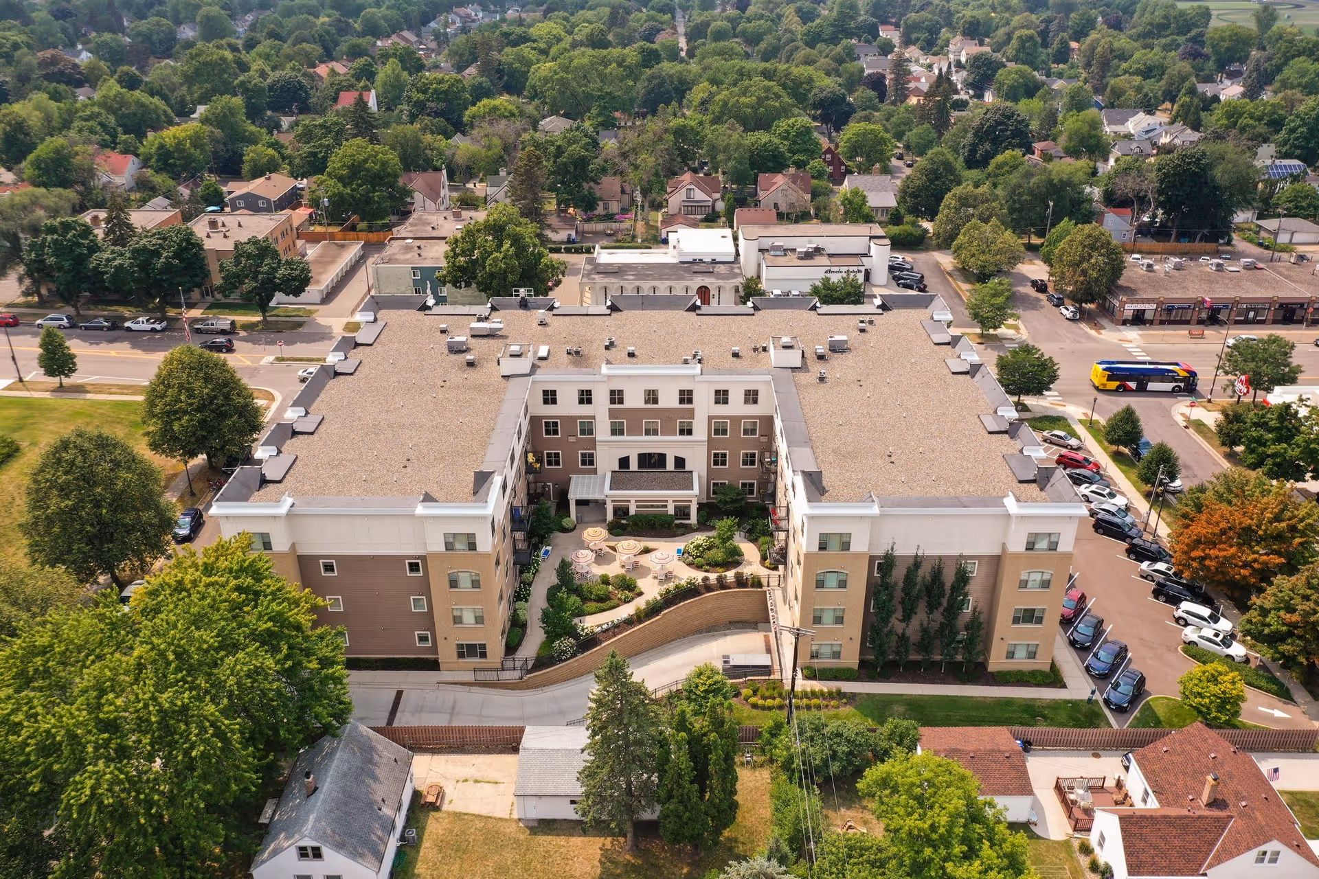 Aerial view of The Waters of Highland Park senior living facility showing a large, U-shaped building with a central courtyard featuring outdoor seating and landscaping. Surrounding the building are trees, residential houses, streets, and parked cars.