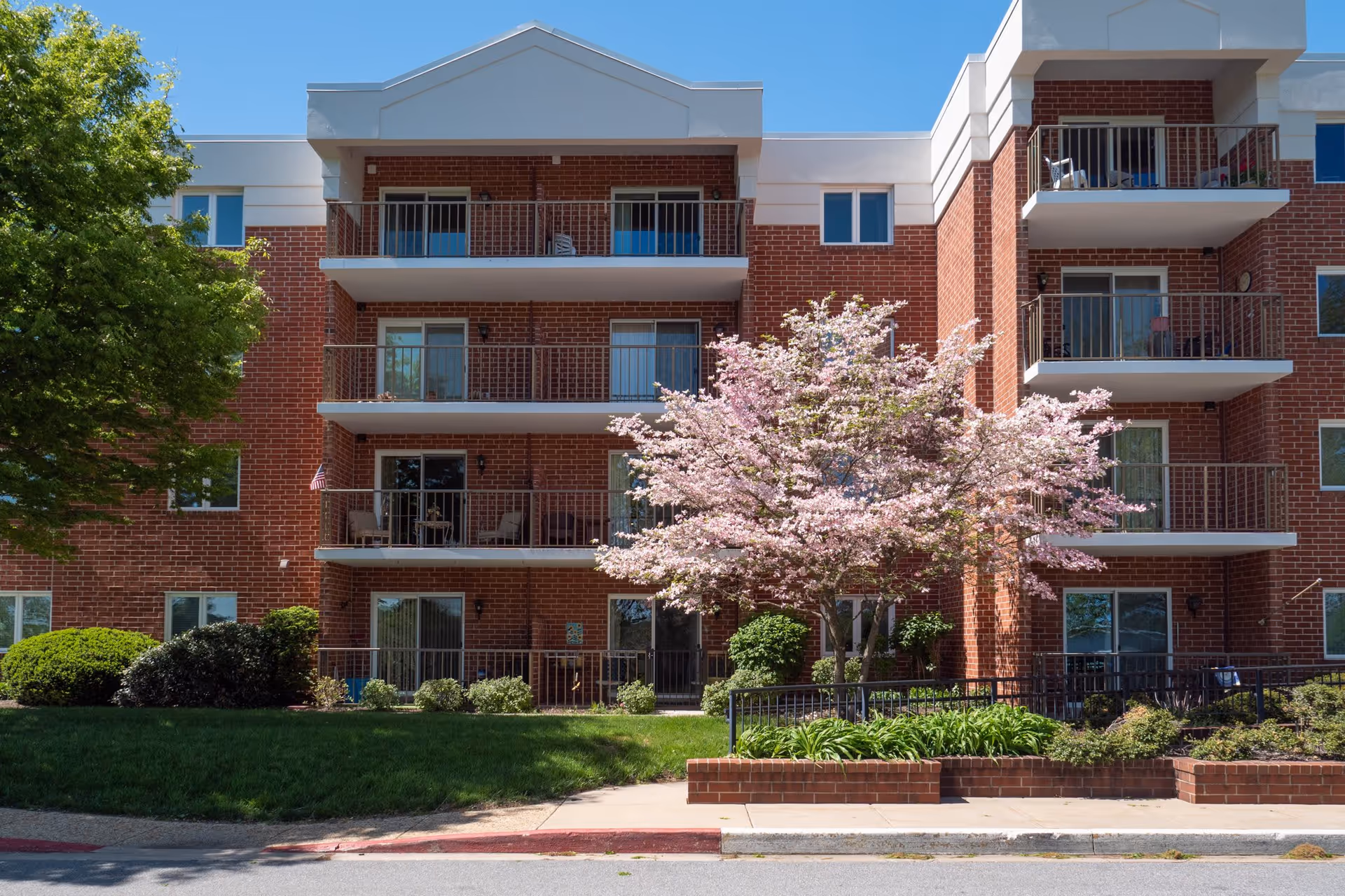 Exterior view of a three-story brick residential building with balconies. In front of the building, there is a sidewalk, a landscaped area with green bushes, and a tree with pink blossoms. The sky is clear and blue.