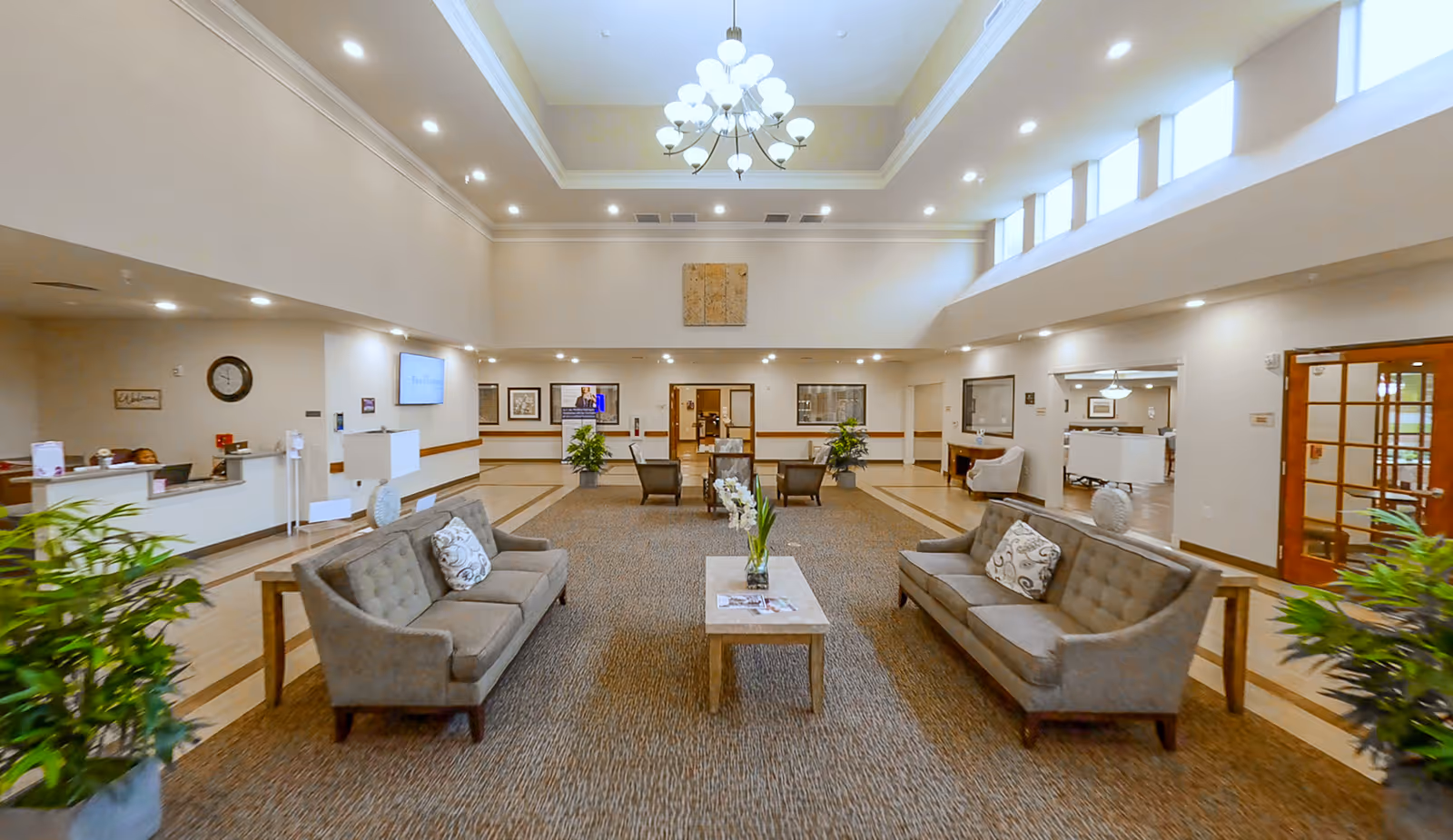 A spacious and well-lit senior living facility common area with two gray sofas facing each other across a coffee table with a flower vase. The room features high ceilings with a chandelier, large windows near the ceiling allowing natural light, and several chairs and plants arranged around the space. The reception desk is visible on the left side, and there are doors and framed pictures on the walls.