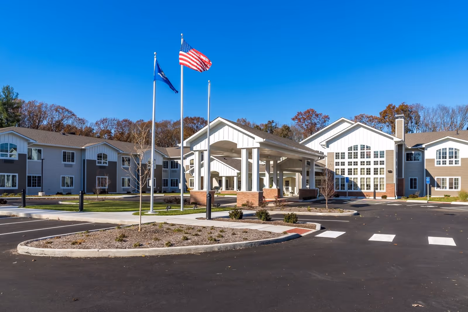 Exterior view of The Landing of North Haven senior living facility showing a large building with white and beige siding, multiple windows, and a covered entrance. Three flagpoles with flags are in front of the building, and there is a paved parking area with landscaped islands under a clear blue sky.