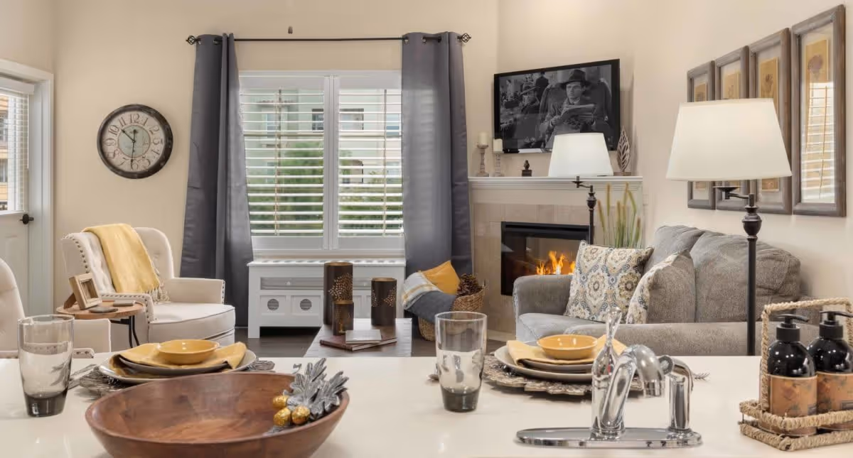 A cozy living room with a gray sofa adorned with patterned pillows, a lit fireplace, and a wall-mounted black and white TV above the mantel. There is a white armchair with a yellow throw blanket and a small side table. A large window with gray curtains and white blinds lets in natural light. In the foreground, a kitchen counter with a wooden bowl, place settings, glasses, and a faucet is visible.