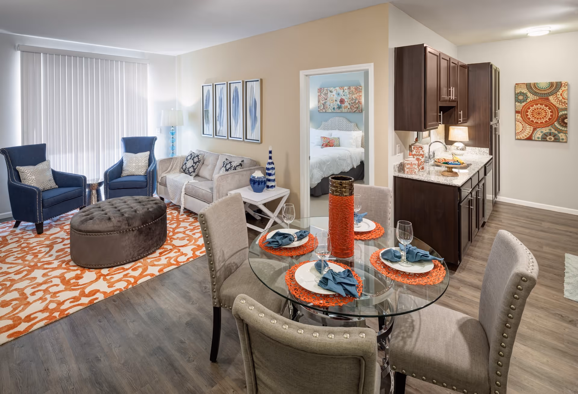 A bright and modern senior living apartment featuring a living room with two blue armchairs, a beige sofa, and a large brown ottoman on an orange patterned rug. Adjacent to the living room is a dining area with a round glass table set for four with orange placemats and blue napkins. The kitchen area has dark wood cabinets and granite countertops. A bedroom with a white bed and colorful artwork is visible through an open doorway.