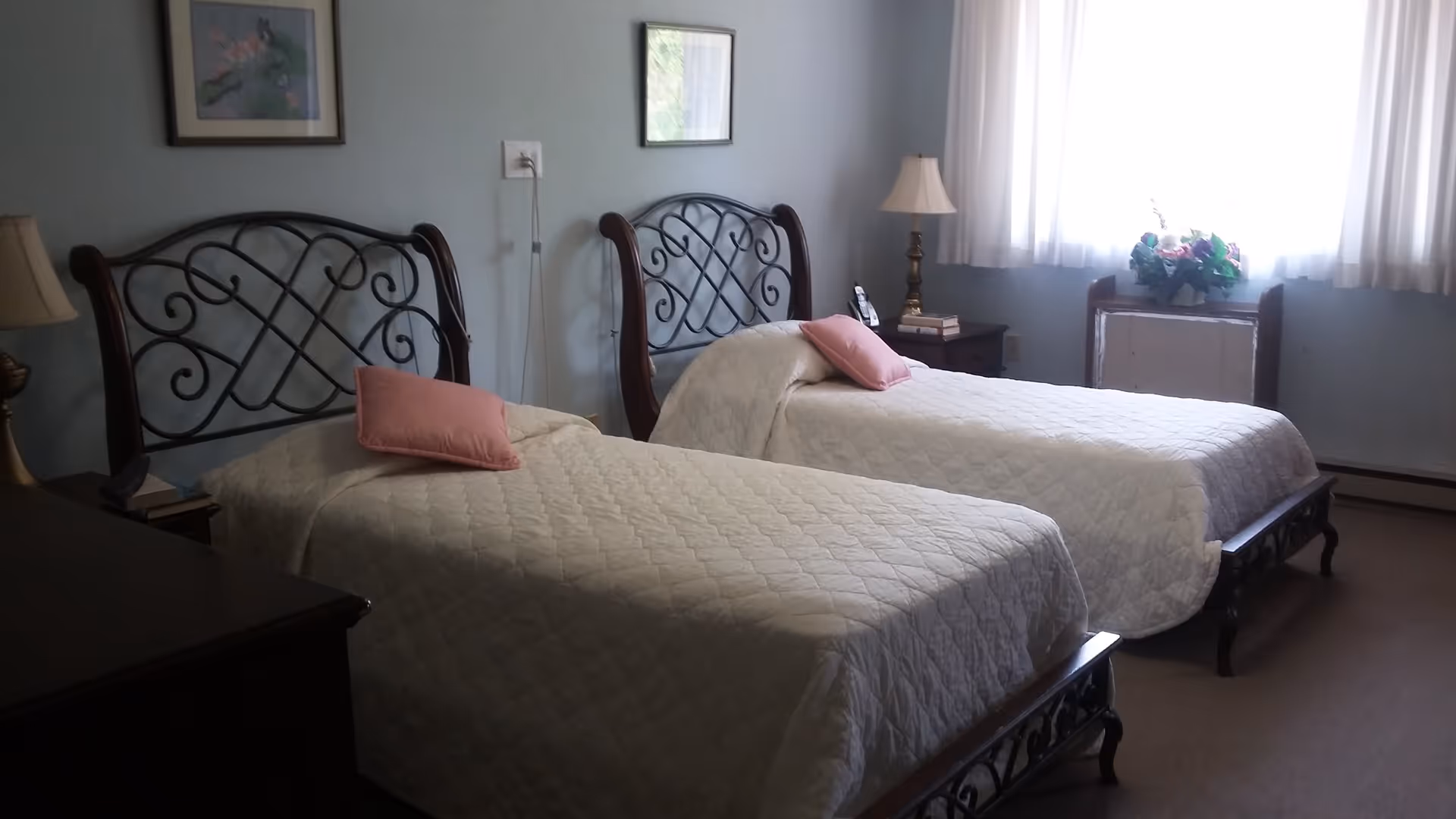 A bedroom with two twin beds featuring ornate metal and wood headboards, each bed covered with a white quilt and a pink pillow. There is a nightstand between the beds with a lamp and books, a window with white curtains letting in natural light, and framed pictures on the light blue walls.