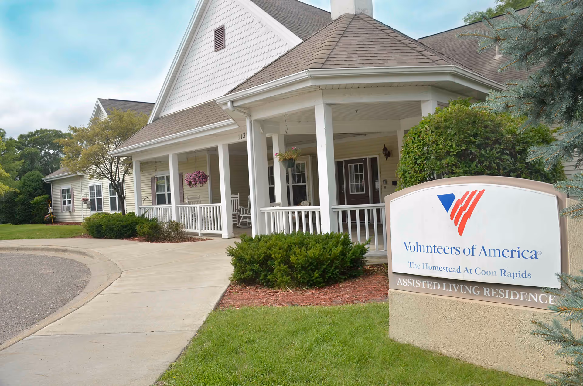 Exterior view of a single-story assisted living residence with a covered porch, white railings, hanging flower baskets, and rocking chairs. A sign in the foreground reads 'Volunteers of America, The Homestead At Coon Rapids, Assisted Living Residence'.