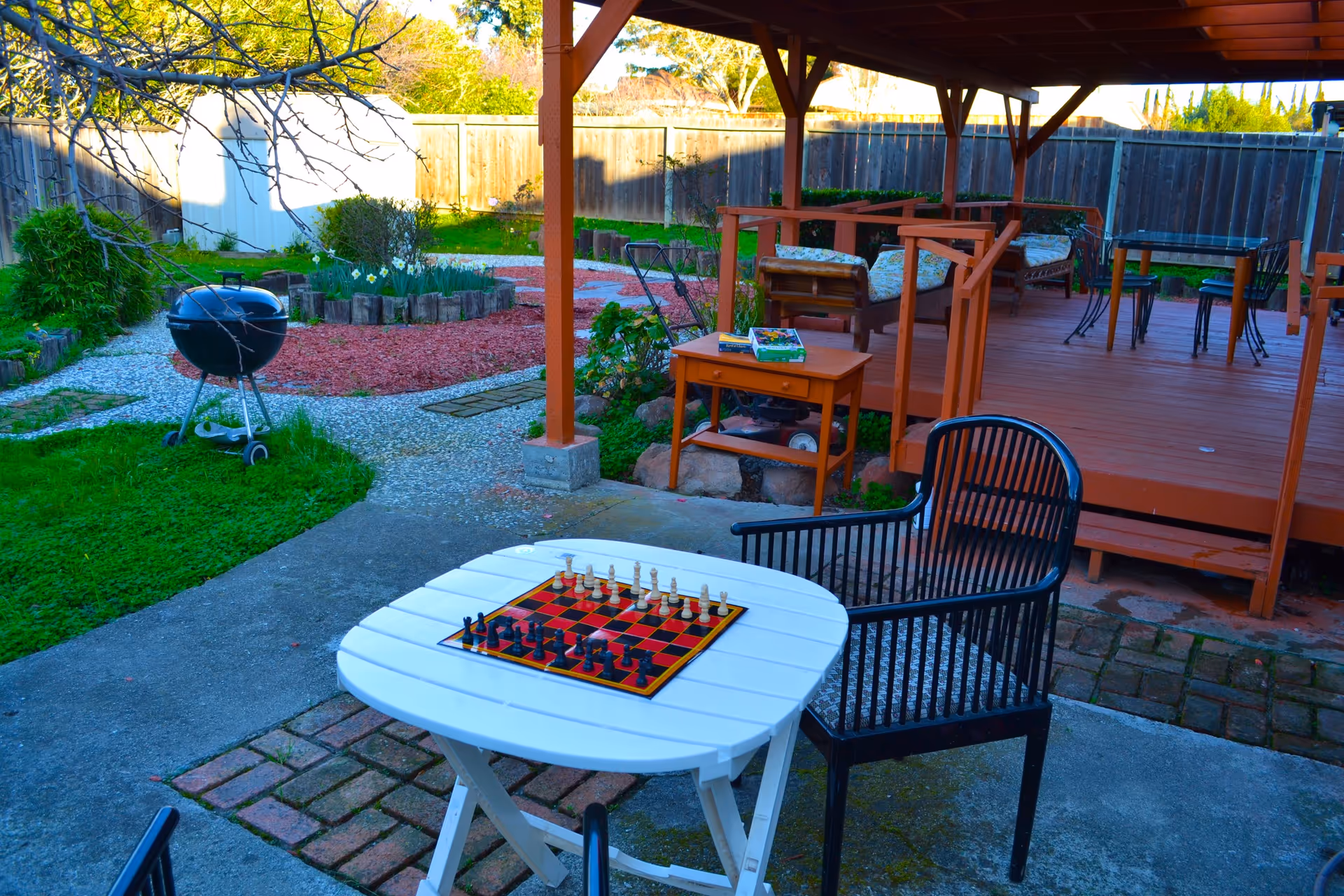 Backyard patio with a covered wooden deck, outdoor seating, a white table with a chessboard, and a charcoal grill.