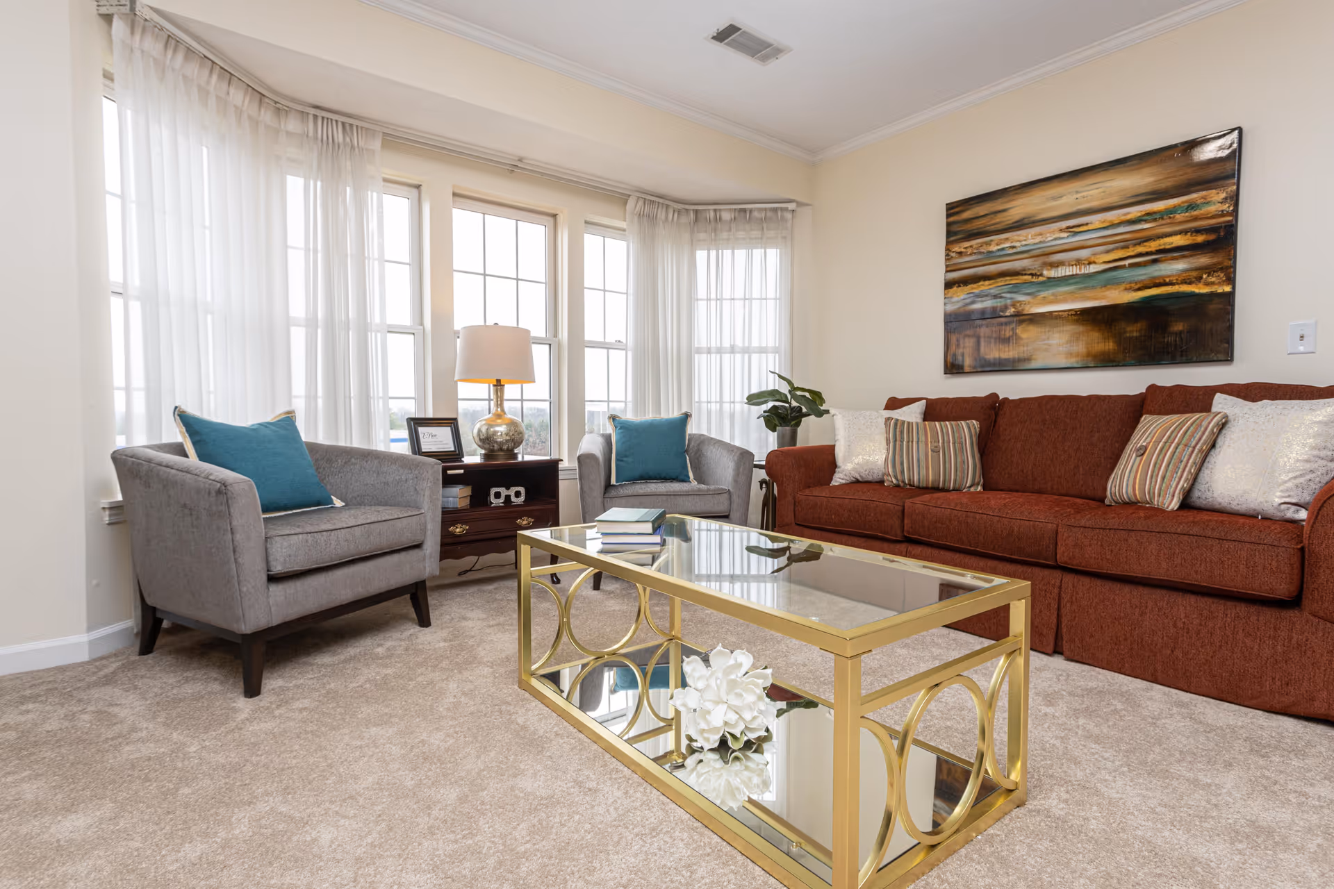 A bright living room with beige carpet, a rust-colored sofa adorned with multiple pillows, two gray armchairs each with a teal cushion, a glass coffee table with a gold frame holding a white flower and some books, a wooden side table with a lamp and framed picture, and large windows with sheer white curtains letting in natural light. A large abstract painting hangs on the wall above the sofa.
