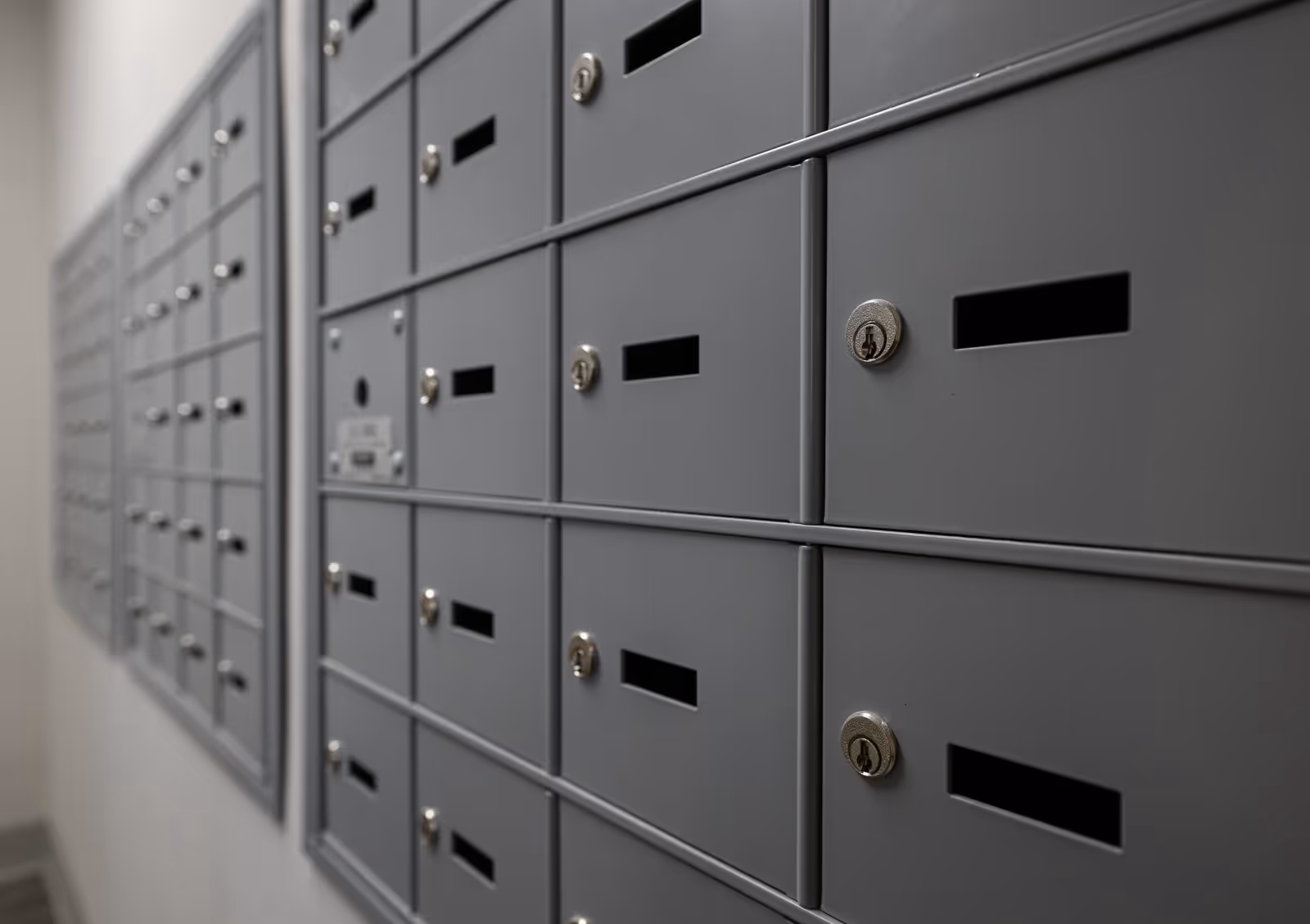 A close-up view of multiple gray metal mailboxes with keyholes and mail slots, mounted on a wall in a hallway.