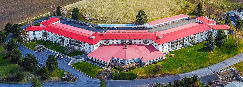 Aerial view of a long, multi-wing building with red roofs set amid lawns, trees, and a curved driveway.