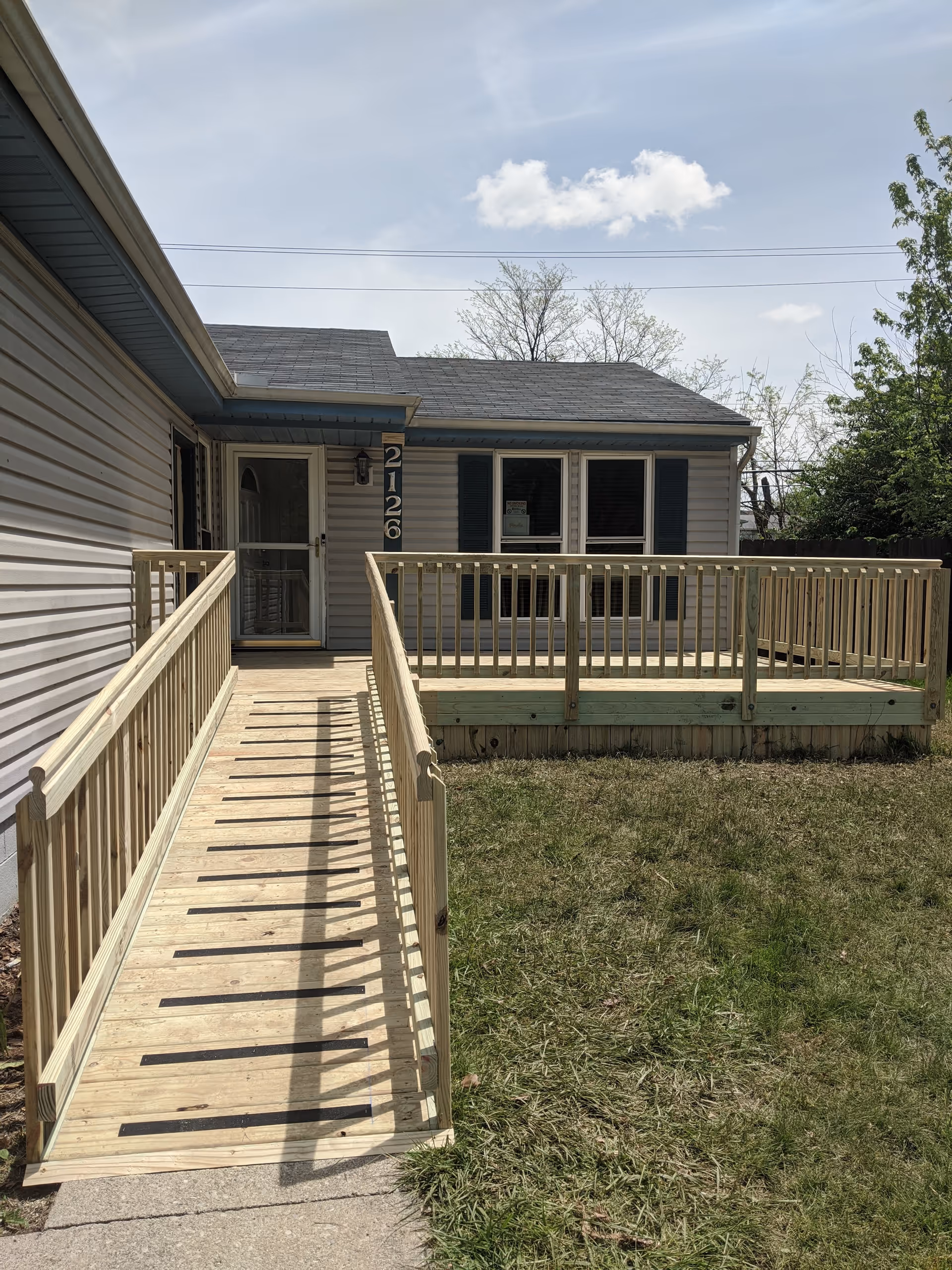 Exterior view of a single-story house with beige siding and a gray shingled roof. There is a wooden wheelchair ramp leading up to the front door, and a wooden deck area to the right of the ramp. The house number 2126 is displayed vertically next to the door. The sky is partly cloudy and there is grass in the foreground.