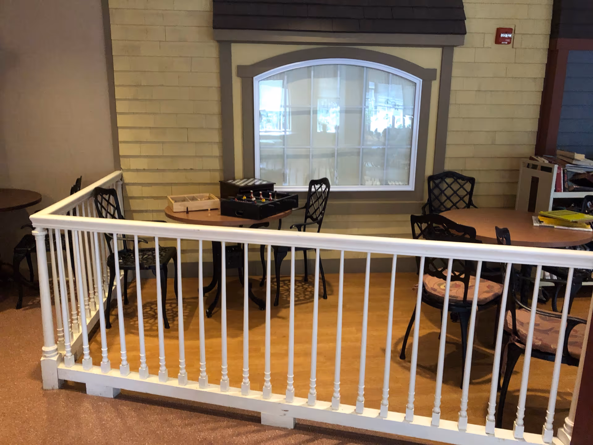 Indoor seating area with round tables and black metal chairs with cushions, a white railing in the foreground, and a window with white curtains on a beige wall in the background. Board games are placed on one of the tables.