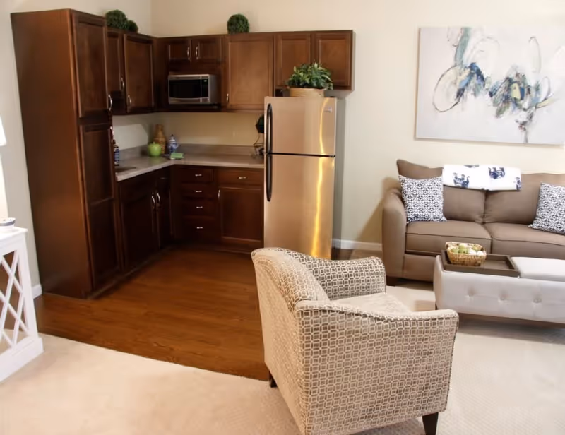 A cozy living area with a beige patterned armchair in the foreground, a brown sofa with decorative pillows and a throw blanket, a tufted ottoman coffee table with a basket of green apples, and a kitchen area with wooden cabinets, a stainless steel refrigerator, and a microwave. A modern abstract painting hangs on the wall above the sofa.
