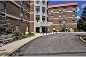 Front entrance of a multi-story brick senior living building with a covered portico, circular driveway, benches, and landscaping.