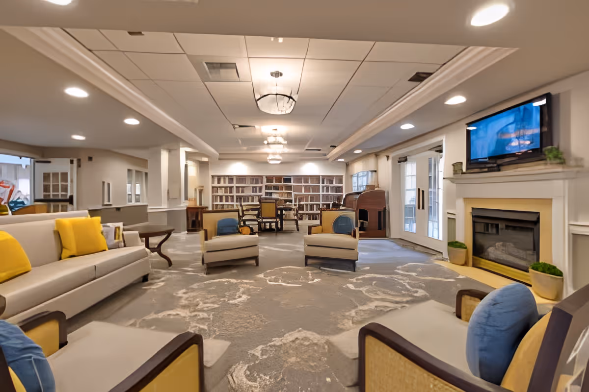 Spacious senior living lounge with sofas and armchairs arranged around a fireplace and TV, with bookshelves at the far end.