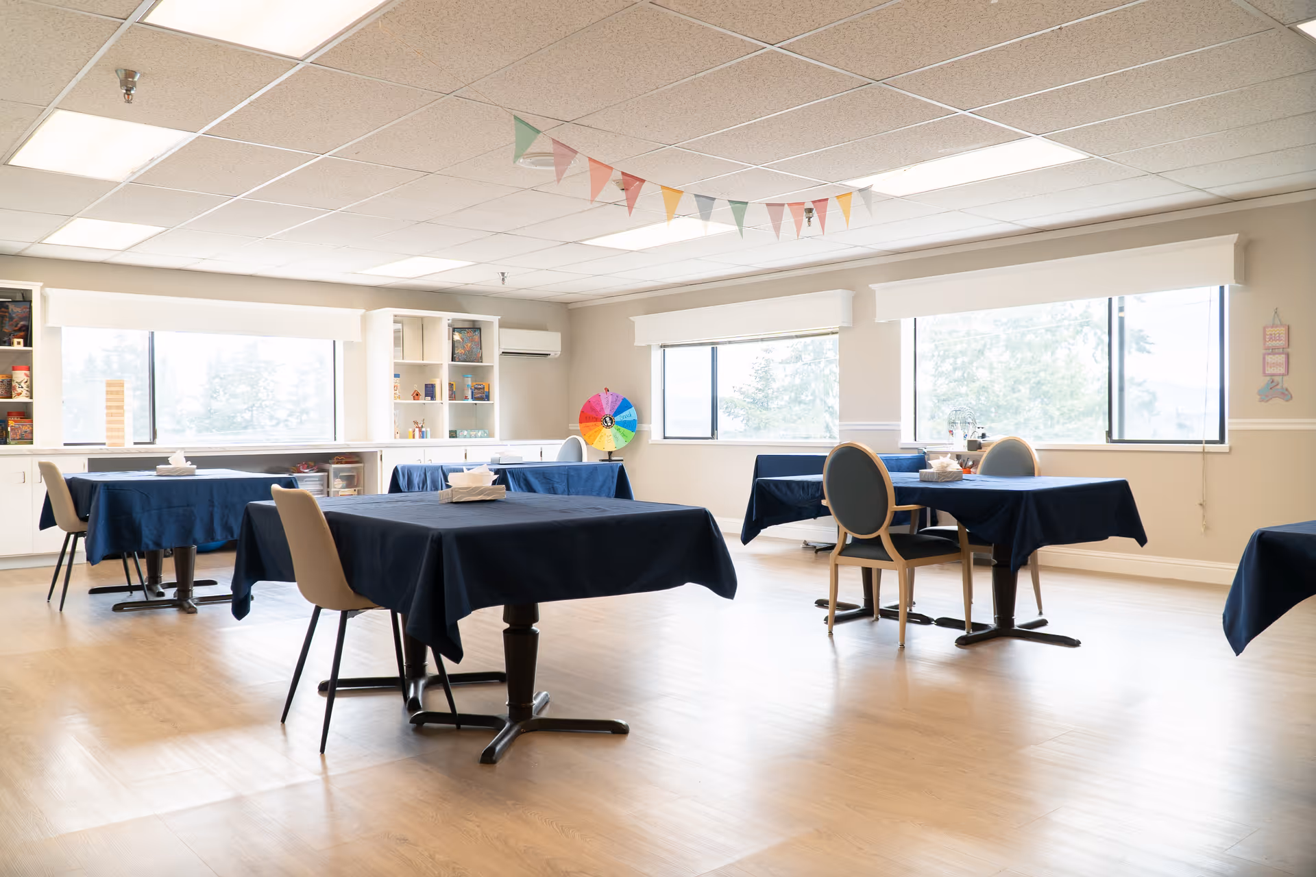 A bright communal dining/activity room with several tables covered in dark tablecloths, chairs, large windows, and decorative bunting.