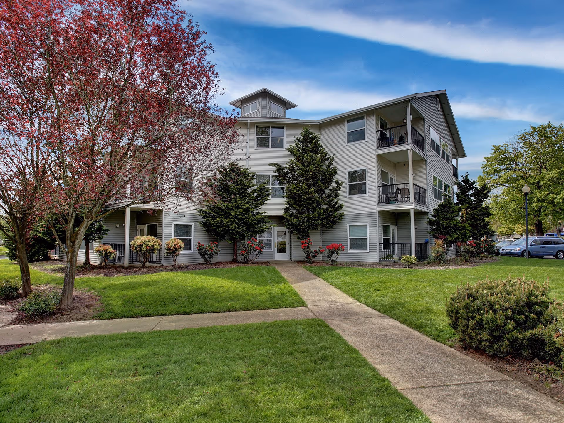 Exterior view of a three-story senior living facility building with balconies, surrounded by green grass, trees with red and green leaves, and a concrete walkway leading to the entrance under a partly cloudy blue sky.