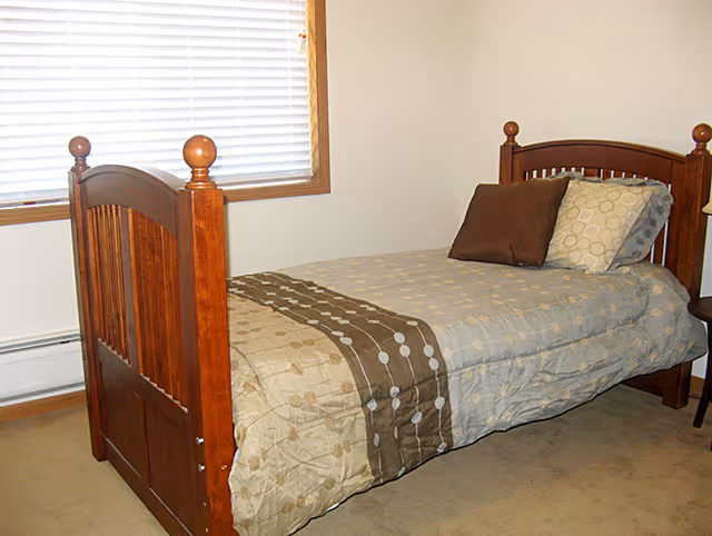A single wooden bed with a headboard and footboard in a bedroom. The bed is made with a patterned comforter in beige and brown tones, and there are three pillows arranged at the head of the bed. A window with closed blinds is visible behind the bed, and the room has beige carpet flooring and light-colored walls.