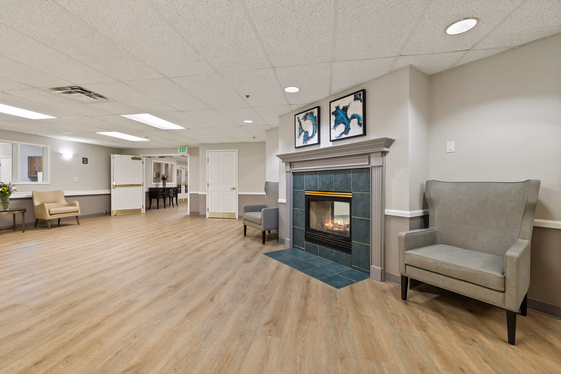 A spacious interior common area with light wood flooring, a fireplace with a tiled surround, two gray armchairs on either side of the fireplace, and two abstract paintings above it. The walls are painted in neutral tones, and there is a beige armchair and a small table with a flower vase in the background near an open door leading to another hallway.
