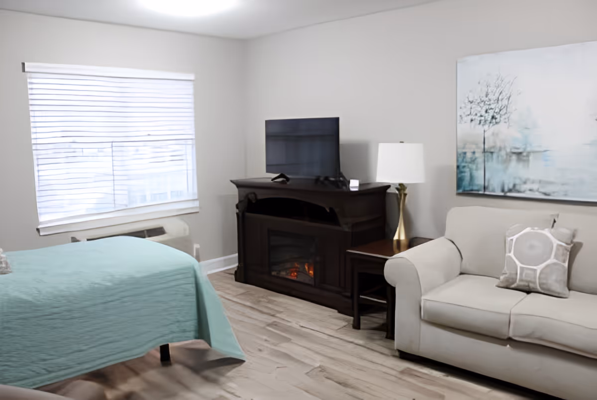 A cozy room with a bed covered in a light blue quilt, a beige sofa with a decorative pillow, a wooden electric fireplace with a TV on top, a side table with a lamp, and a large window with blinds letting in natural light. A painting with a tree and water scene hangs on the wall above the sofa.