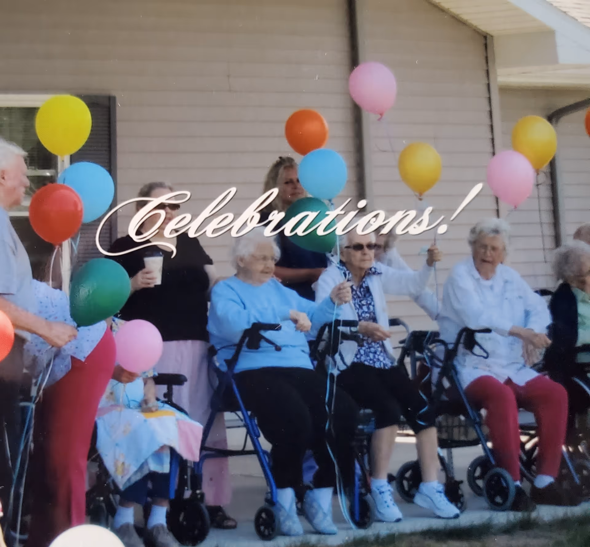 A group of elderly people sitting outside in front of a building, holding colorful balloons. Some are seated in wheelchairs or using walkers. The word 'Celebrations!' is overlaid on the image.