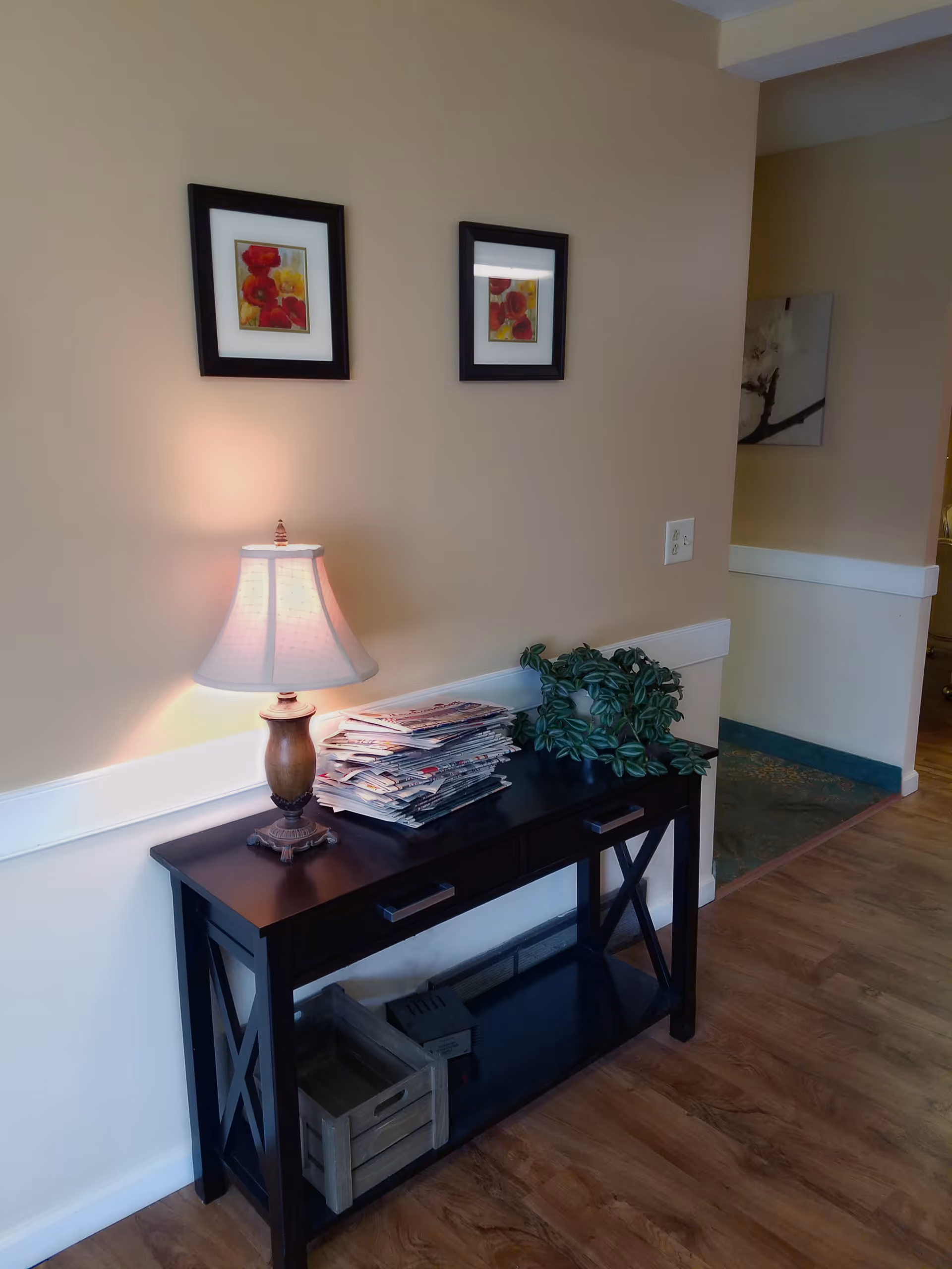 A hallway console table against a beige wall holds a lamp, a stack of newspapers, and a potted plant beneath two framed floral prints.