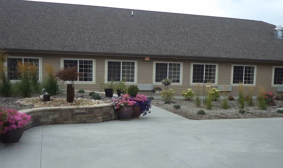 Exterior view of a single-story building with multiple windows, a sloped roof, and a landscaped garden area featuring various plants and flowers in pots and beds. The foreground shows a concrete pathway or driveway.