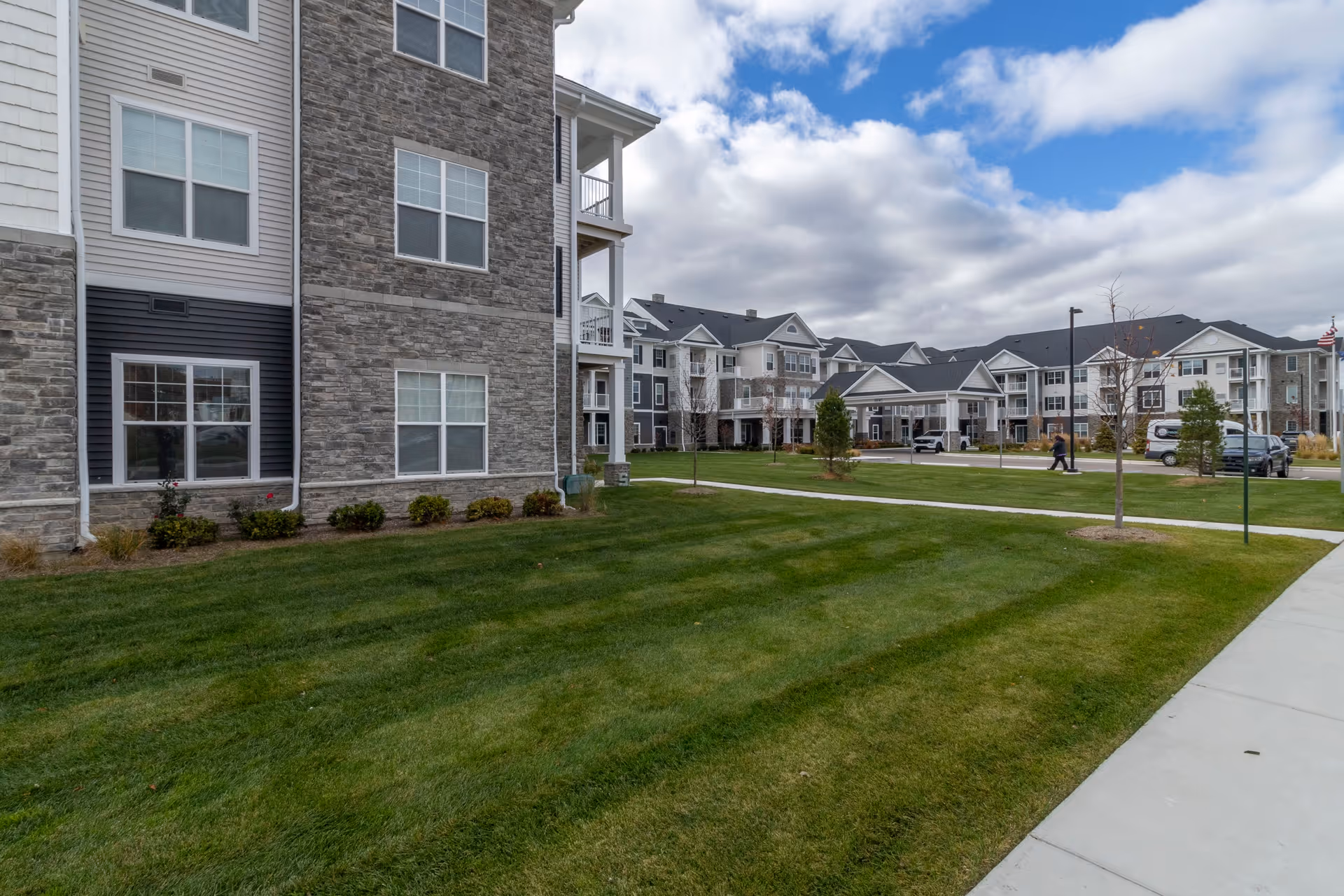 Exterior view of StoryPoint Clinton Township senior living facility showing multiple connected buildings with stone and siding facades, well-maintained green lawns, a sidewalk, and a partly cloudy sky.