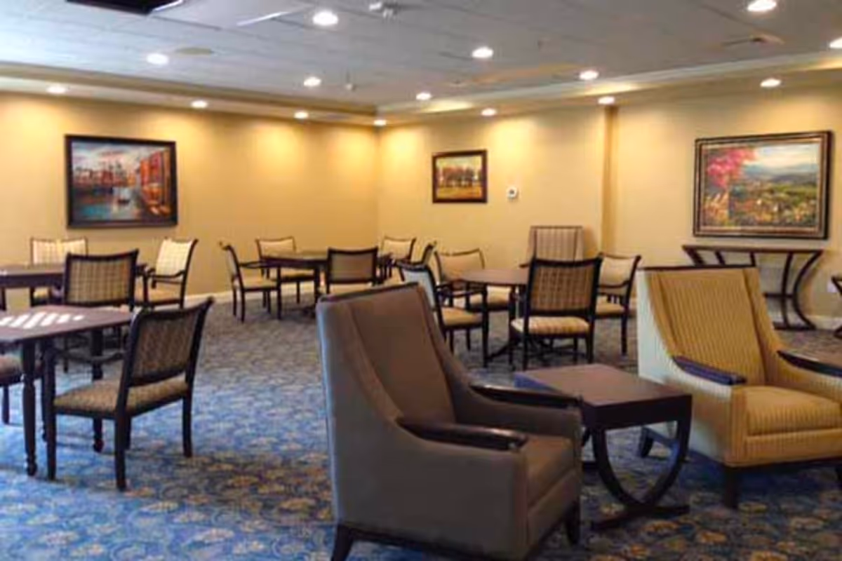 A bright communal dining and lounge area with multiple tables, chairs, and armchairs in a senior living facility.