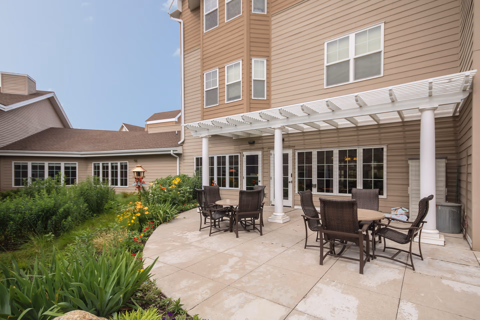 Outdoor patio area at The Deerfield with round tables and wicker chairs under a white pergola attached to a beige building. There are green plants and flowers along the edge of the patio.