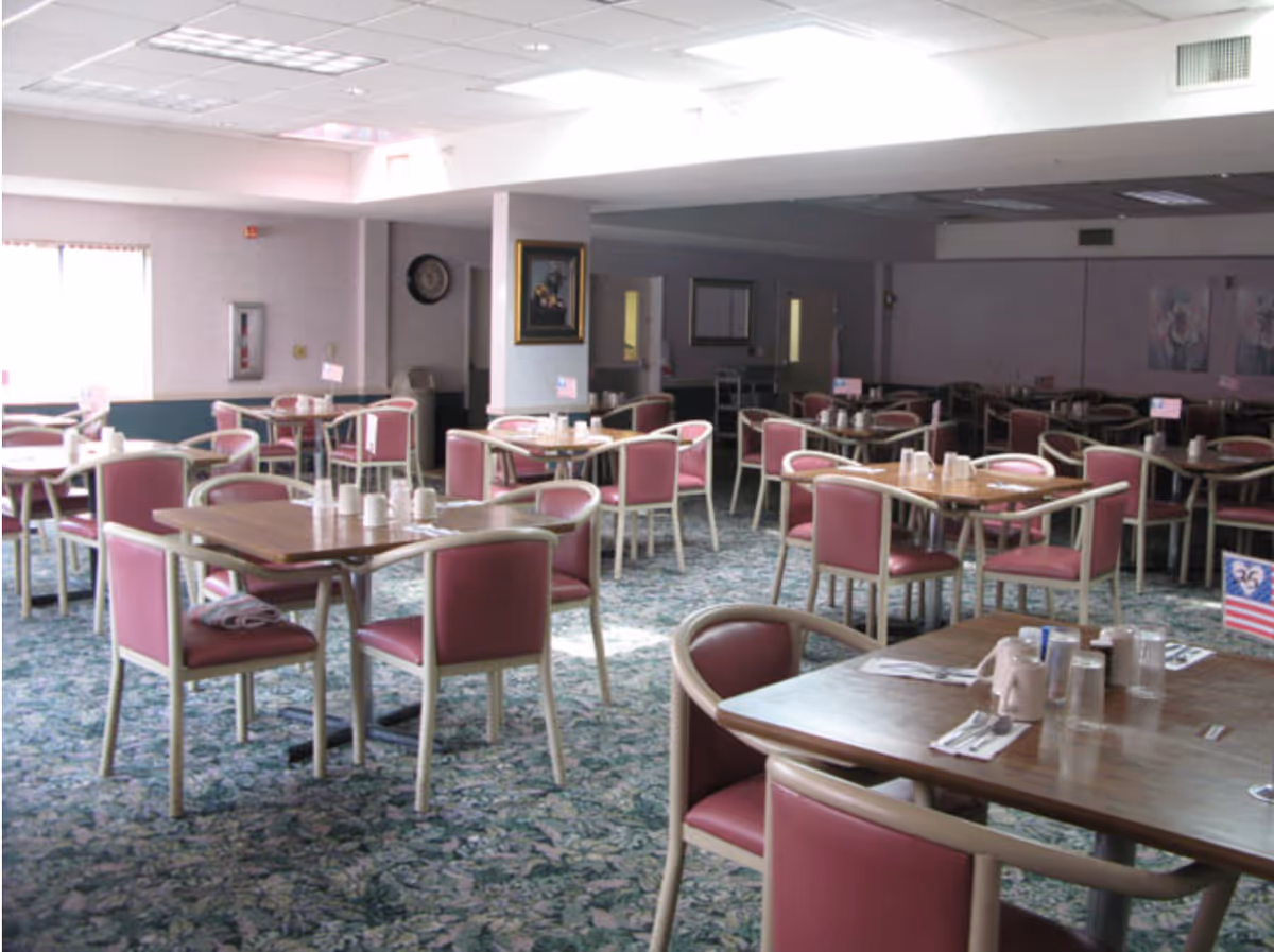 A senior living community dining room with multiple wooden tables and pink cushioned chairs arranged neatly. Each table is set with cups, utensils, and napkins. The room has a patterned carpet, soft lighting, and framed artwork on the walls.