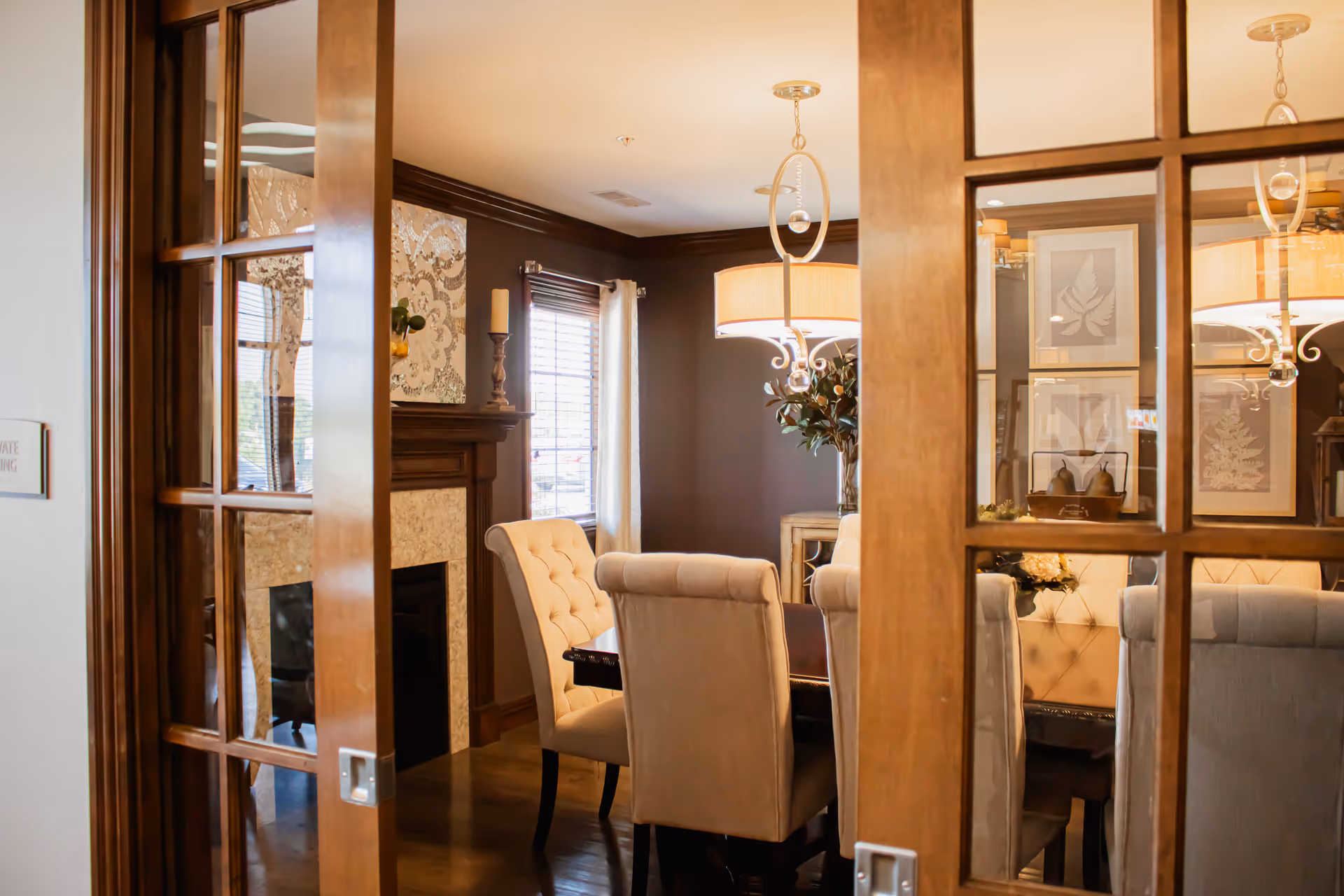 View through wooden French doors into an elegant dining room with tufted chairs, a chandelier, and a fireplace.