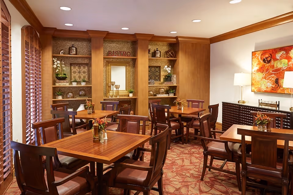 A cozy dining room with wooden tables and chairs arranged neatly. The room features built-in wooden shelves decorated with plants, vases, and a mirror. The word 'BISTRO' is displayed on the central shelf. There are large windows with wooden blinds on the left side, a patterned carpet, and a colorful abstract painting on the right wall above a dark wooden sideboard with two lamps.