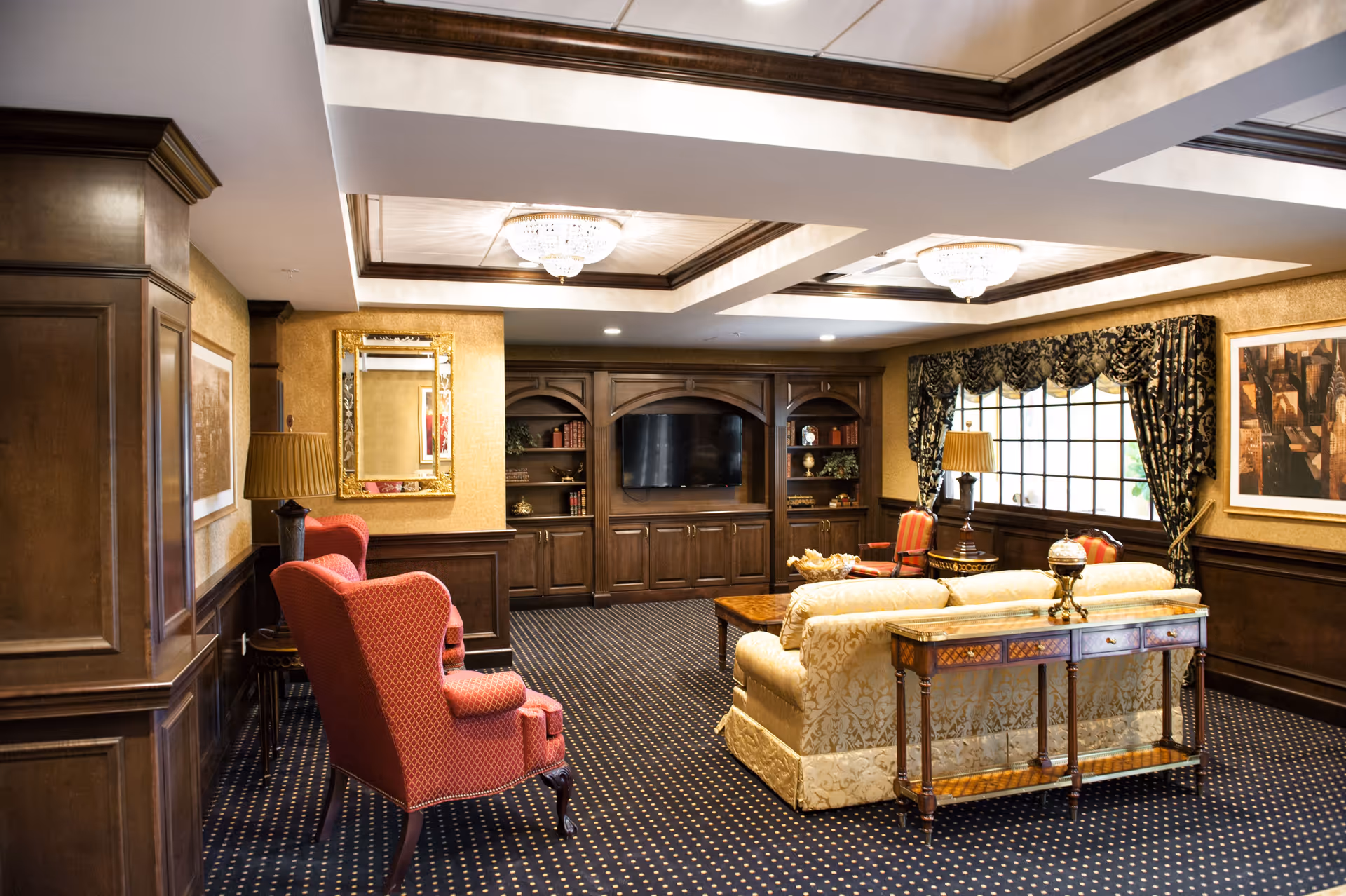 A cozy living room area with classic decor featuring a beige patterned sofa, two red upholstered armchairs, a wooden coffee table, and a wooden console table with decorative items. The room has dark wood paneling, a built-in wooden entertainment center with a flat-screen TV, patterned carpet, and large windows with dark patterned curtains. Two elegant chandeliers hang from the ceiling.