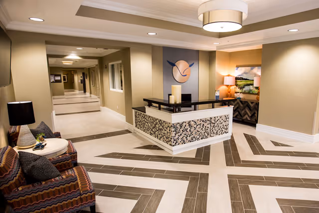 A modern and well-lit reception area in an assisted living facility featuring a decorative front desk with candles, patterned flooring, a seating area with two colorful armchairs and a small round table with a lamp, and a hallway leading to other rooms.