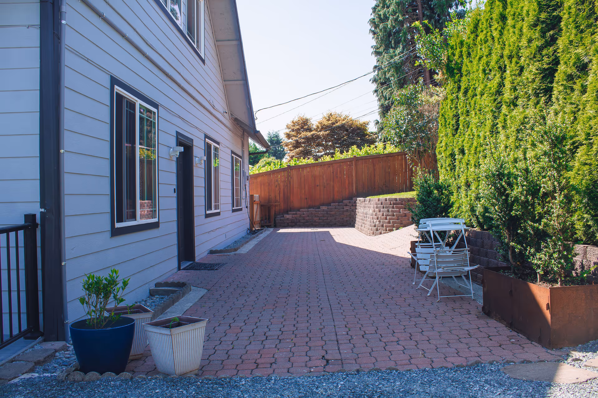 Paved side yard of a light-gray house with potted plants, folding chairs, tall hedges, and a wooden fence.
