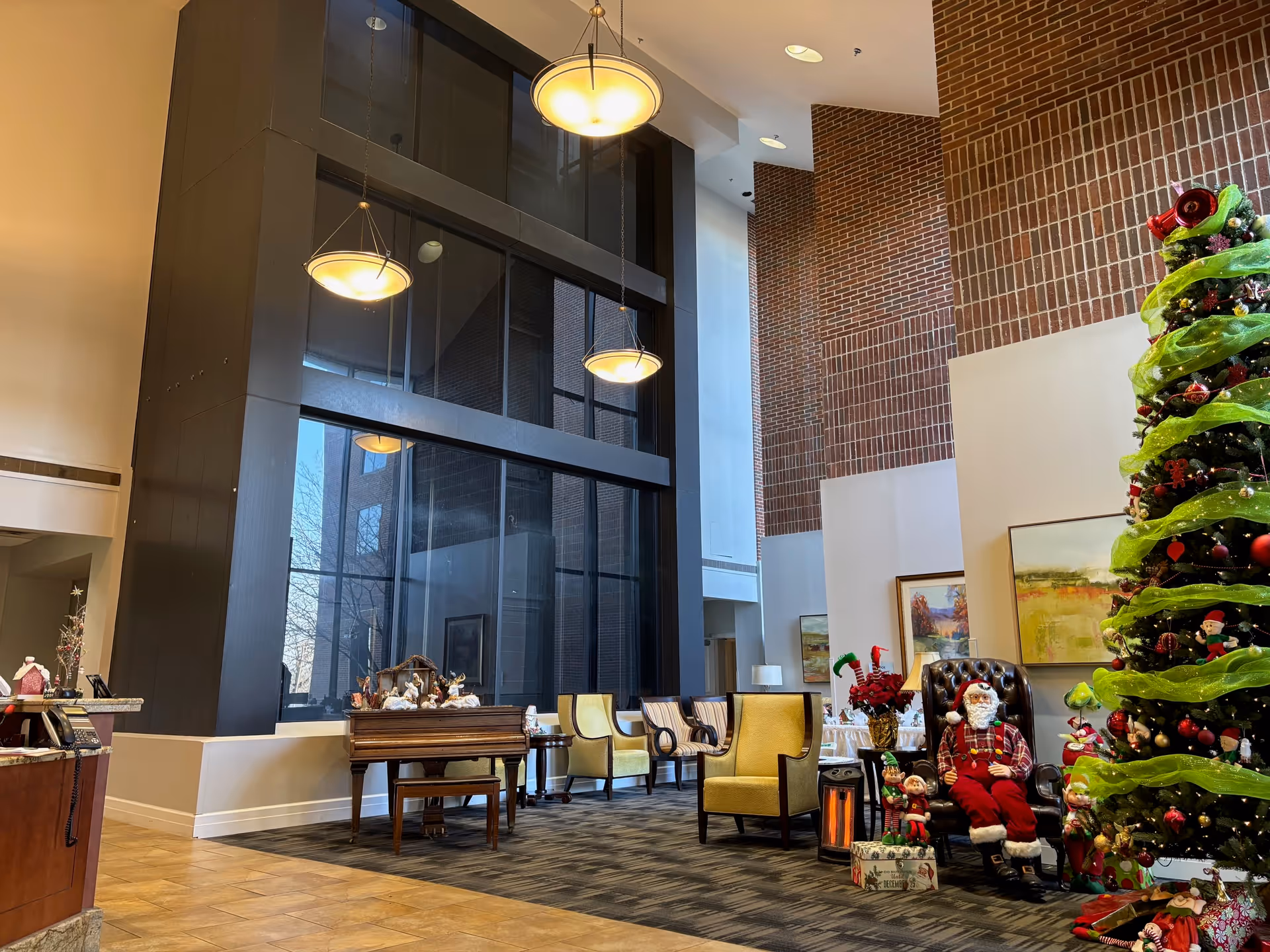 A spacious and well-lit lobby area with high ceilings and large windows. The room features a decorated Christmas tree with green ribbon and ornaments, a Santa Claus figure seated in a leather chair, and several elf decorations. There are multiple chairs arranged along the wall, a piano with a nativity scene on top, and hanging pendant lights illuminating the space. The walls are a mix of brick and white surfaces.
