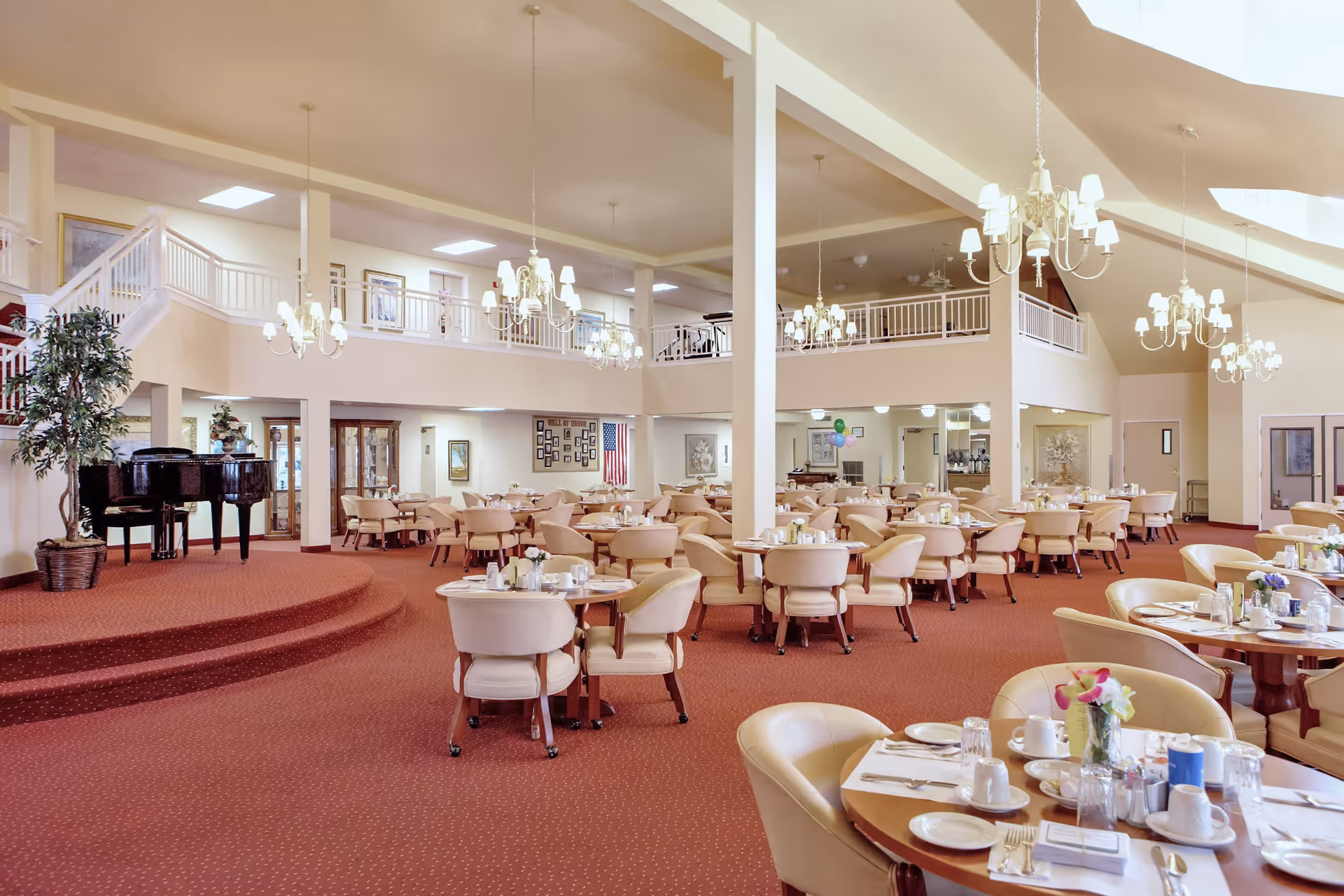 Spacious dining room with multiple round tables set with white cups, plates, and silverware. Cream-colored cushioned chairs surround the tables. The room features red carpet flooring, cream walls, and several chandeliers hanging from the ceiling. A black grand piano is positioned on a small raised platform to the left, and there is a second-floor balcony overlooking the dining area.