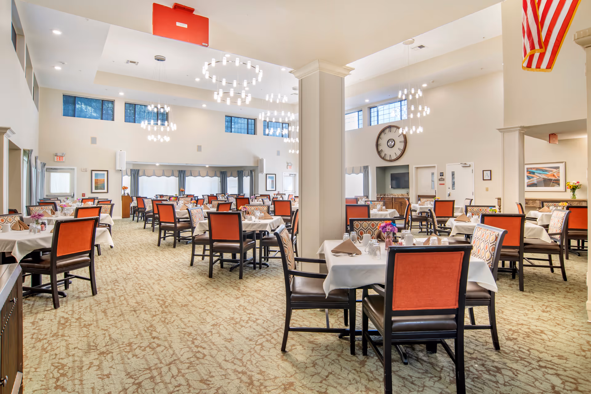 Spacious dining hall with white tablecloths, red chairs, chandeliers, and an American flag.