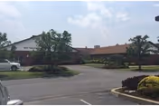 Single-story brick health care building with a covered entrance, circular driveway, parked cars and landscaped grounds under a partly cloudy sky.