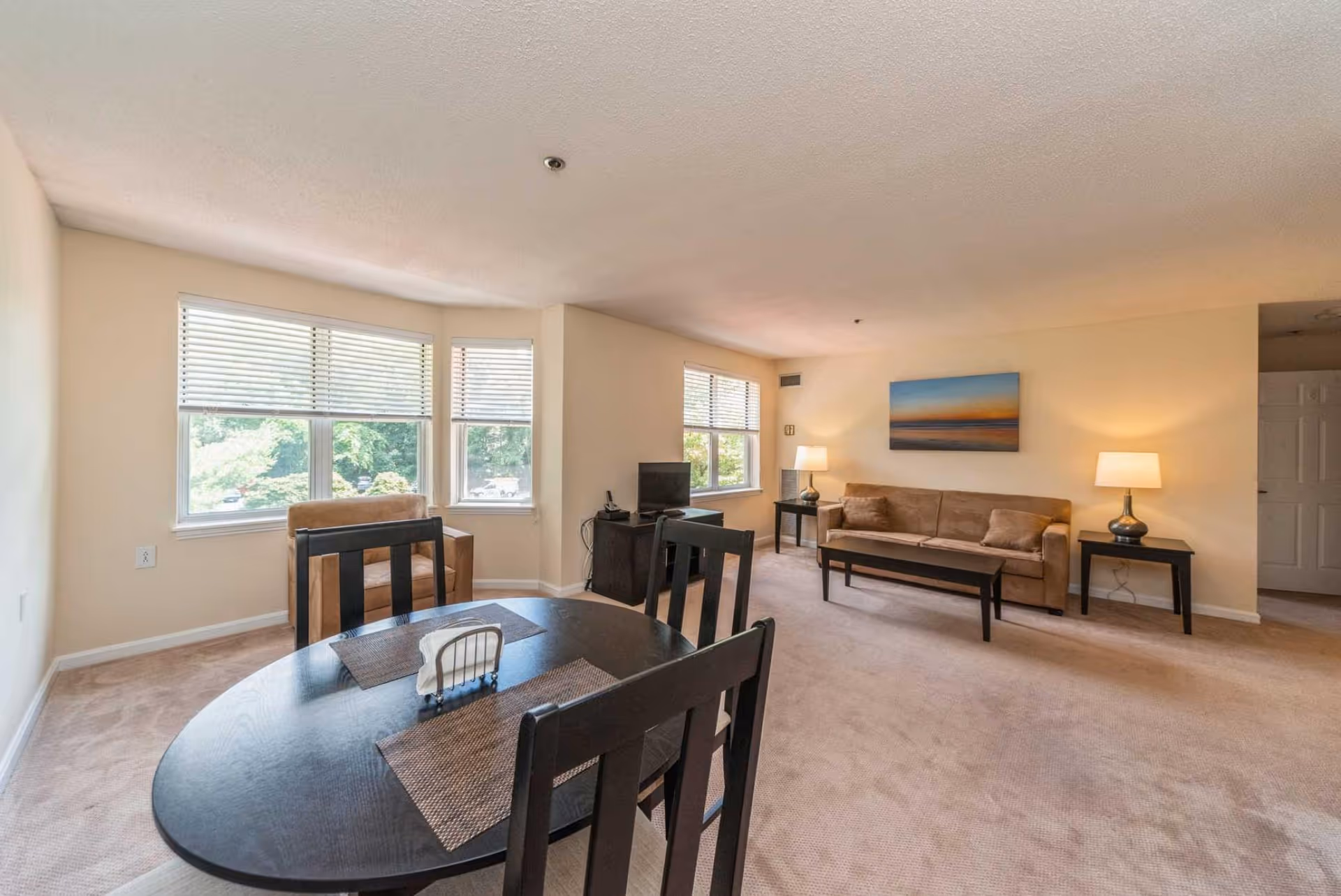 Open living and dining room with a round dining table and chairs in the foreground, a sofa and coffee table near windows and lamps along the far wall.