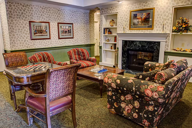 A cozy living room area with floral and striped upholstered chairs and sofa arranged around a wooden coffee table with a chess set. The room features a fireplace with a black marble surround, built-in white shelves with decorative items, patterned wallpaper, and framed artwork on the walls.