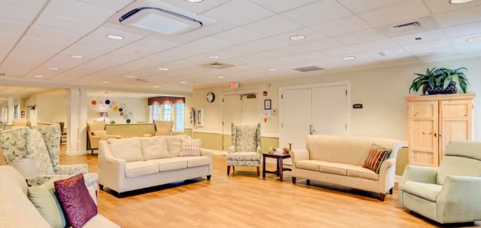 A spacious and well-lit living room area in a senior living facility with multiple beige and patterned armchairs and sofas arranged around small tables on a wooden floor. The walls are light-colored, and there is a wooden cabinet with a plant on top. The ceiling has recessed lighting and air vents.