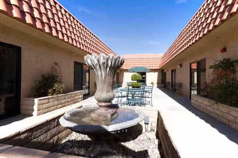 Sunny courtyard with a central fountain, planters, and patio seating between beige buildings.