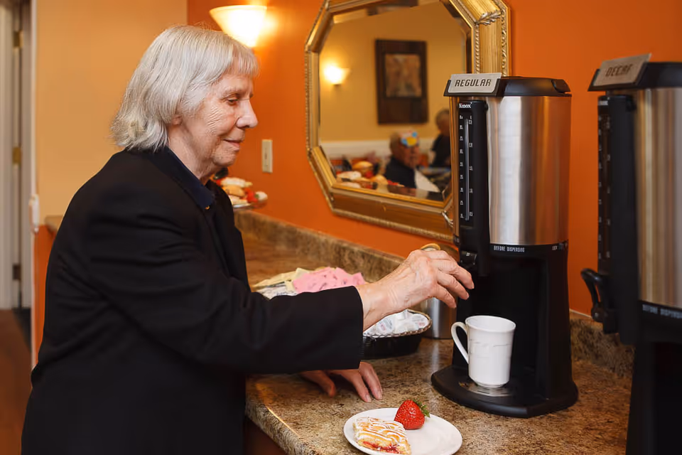 An elderly woman with gray hair is dispensing coffee from a coffee machine labeled 'Regular' into a white mug on a countertop. Next to the mug is a plate with a pastry and a strawberry. The background shows a mirror reflecting part of the room with warm lighting and another coffee machine labeled 'Decaf'.