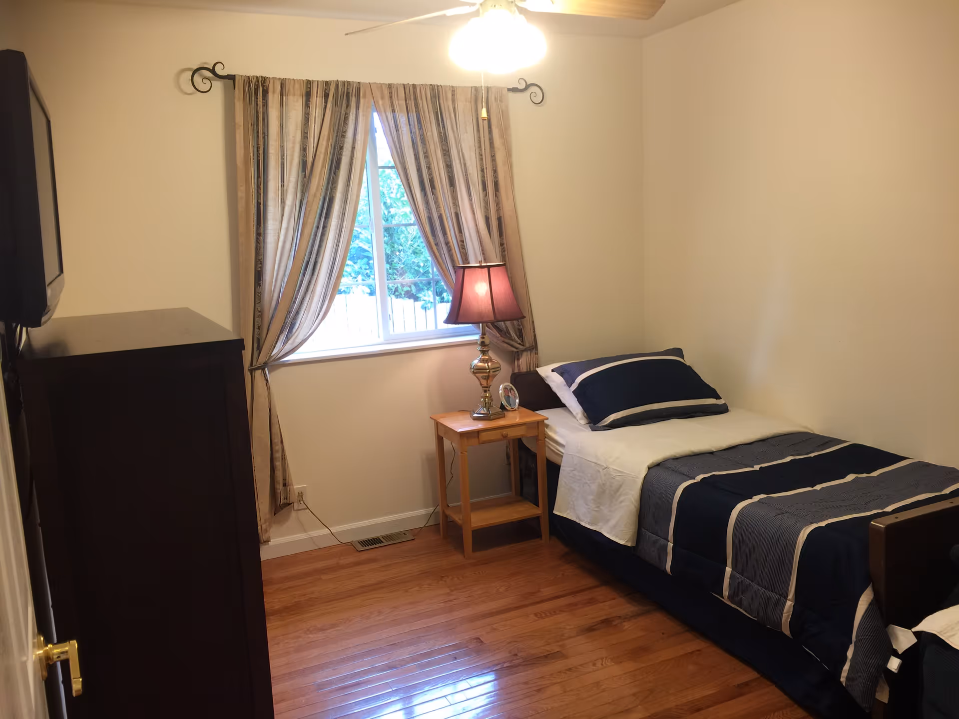 A small bedroom with a single bed made with navy blue and white bedding. There is a wooden nightstand next to the bed with a lamp and a small clock. A window with patterned curtains is behind the nightstand. A dark wooden dresser is positioned against the left wall, and the floor is hardwood.
