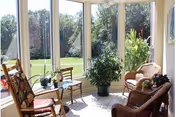Sunlit sunroom with wicker chairs, potted plants, and large windows overlooking a grassy garden.